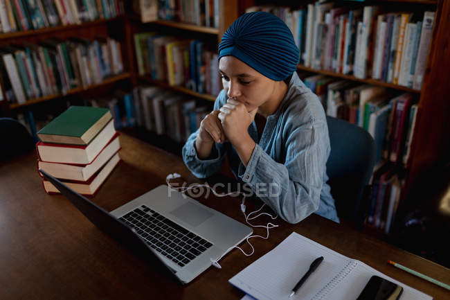 Vista lateral de cerca de una joven estudiante asiática usando un turbante usando una computadora portátil y estudiando en una biblioteca - foto de stock