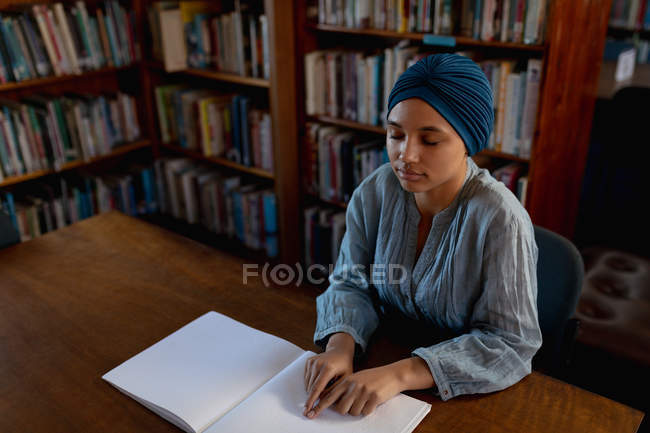 Vista lateral de una joven estudiante asiática ciega usando un turbante leyendo Braille y estudiando en una biblioteca - foto de stock