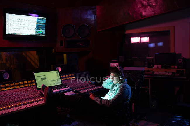 Вигляд молодого кавказького звукоінженера сидить і працює за столом змішування в студії за допомогою ноутбука і носить навушники. — стокове фото