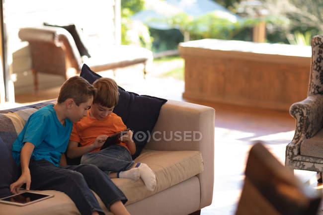Vista lateral de cerca de dos niños caucásicos pre adolescentes sentados en un sofá y utilizando tabletas en la sala de estar - foto de stock