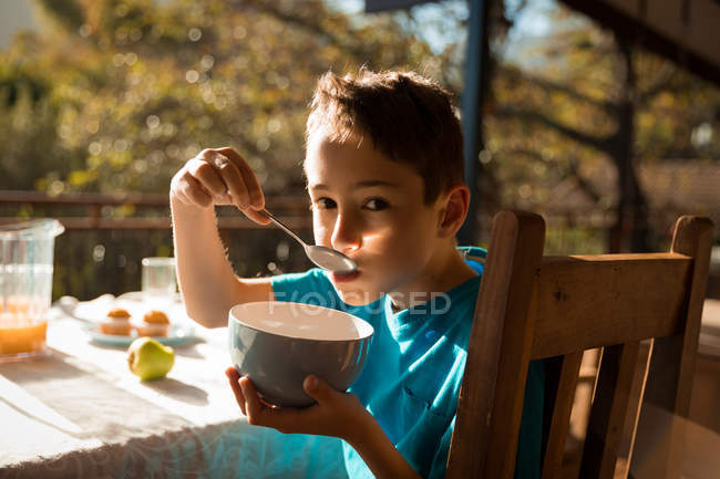 Retrato de un niño caucásico preadolescente sentado en una mesa disfrutando del desayuno en un jardín, comiendo de un tazón - foto de stock