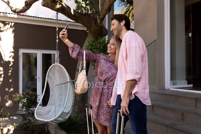 Side view of a happy young Caucasian couple relaxing on holiday standing with suitcases outside a hotel taking a selfie — Stock Photo