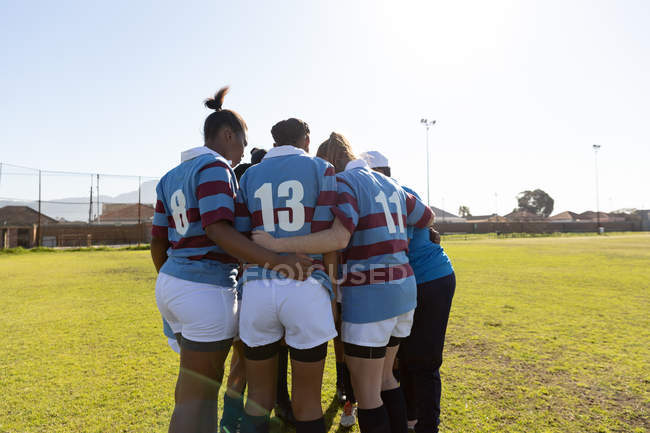 Veduta posteriore di una squadra di giovani giocatrici di rugby multietniche adulte e del loro allenatore in piedi in un raduno su un campo di rugby che si prepara per una partita di rugby — Foto stock