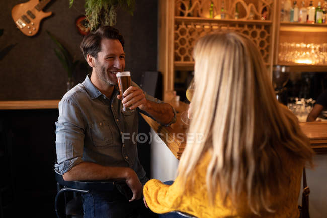 Vista frontal de una feliz pareja de jóvenes caucásicos relajarse juntos en vacaciones en un bar, beber cerveza, vino y sonreír - foto de stock