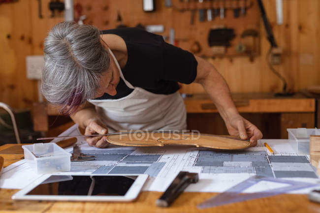 Front view of a senior Caucasian female luthier measuring the body of a violin on a workbench in her workshop, with a tablet computer in front of her and tools hanging up on the wall in the background — Stock Photo