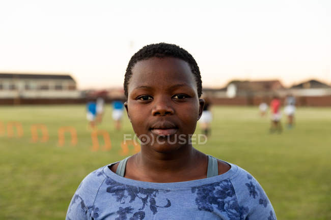 Portrait close up of a young adult African American female rugby player standing on a sports field looking to camera, with her teammates in the background — Stock Photo