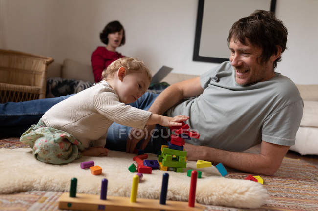 Передній вигляд нагадує молодого кавказького батька, який грає з дитиною на підлозі, а молода кавказька мати сидить на задньому плані. — стокове фото
