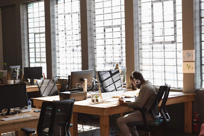 Vista lateral de un joven caucásico sentado en un escritorio junto a una ventana usando un teléfono inteligente y usando auriculares en una oficina creativa - foto de stock