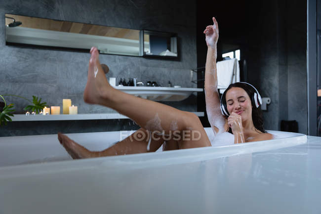 Front view of a young Caucasian brunette woman sitting in a foam bath wearing headphones, listening to music with her eyes closed and arm raised in the air — Stock Photo
