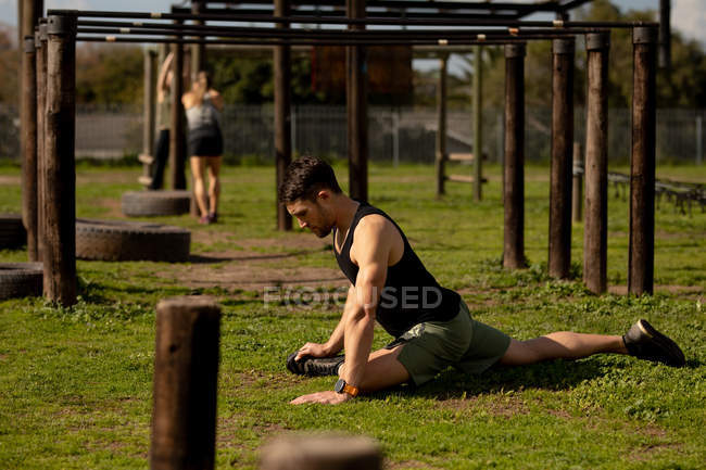 Vista lateral de un joven hombre caucásico sentado en la hierba estirándose en un gimnasio al aire libre antes de una sesión de entrenamiento de bootcamp - foto de stock