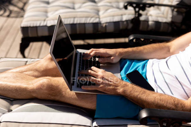 Вид збоку посередині чоловіка, який відпочиває у відпустці, сидить на сонячному ліжку і використовує ноутбук — стокове фото