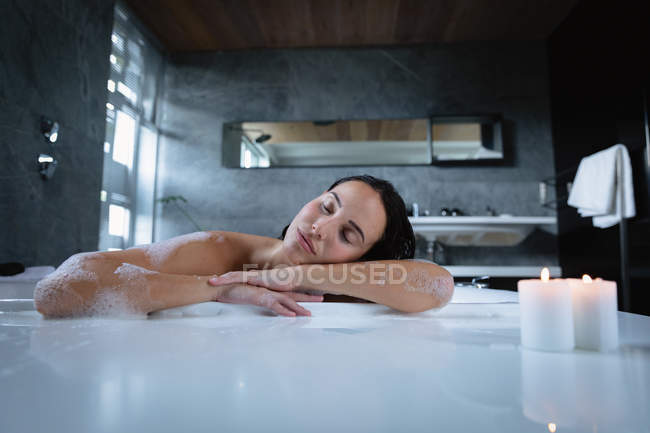 Портрет молодої кавказької брюнеткою, яка сидить у пінній ванні з запалені свічками на краю, спираючись збоку і відпочиває з закритими очима — стокове фото