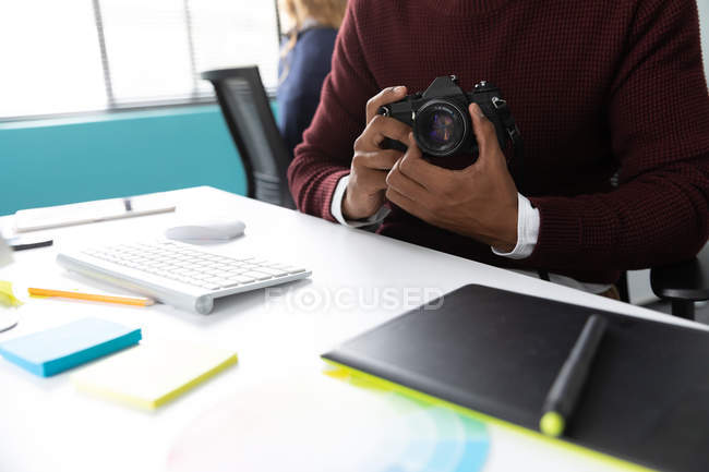 Front view mid section of man sitting at a desk holding an SLR camera in the modern office of a creative business — Stock Photo