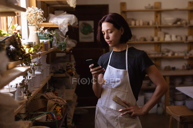 Vista frontale una giovane ceramista caucasica in piedi con la mano sul fianco usando uno smartphone in uno studio di ceramica — Foto stock