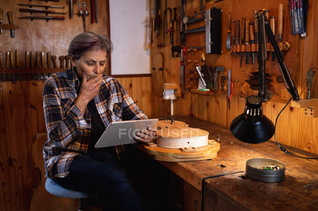 Front view of a senior Caucasian female luthier using a tablet computer with tools hanging up on the wall in the background — Stock Photo