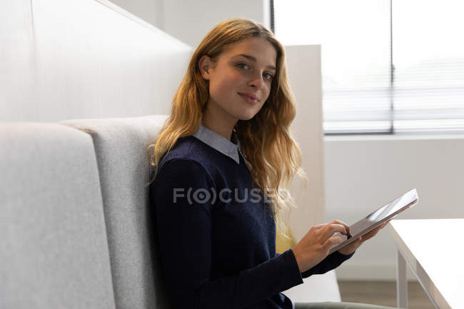 Vista lateral de una joven mujer caucásica sentada en un asiento de banco usando una tableta en el comedor de un negocio creativo, girando y sonriendo a la cámara - foto de stock