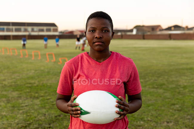 Retrato de una joven jugadora de rugby afroamericana adulta parada en un campo de deportes sosteniendo una pelota de rugby y mirando a la cámara, con sus compañeras de equipo en el fondo - foto de stock