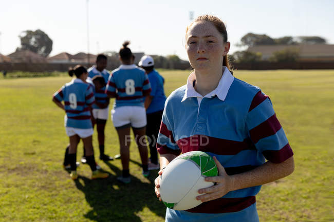 Retrato de uma jovem adulta caucasiana jogadora de rugby em pé em um campo de rugby segurando uma bola de rugby em suas mãos olhando para a câmera, com seus companheiros de equipe conversando juntos no fundo — Fotografia de Stock