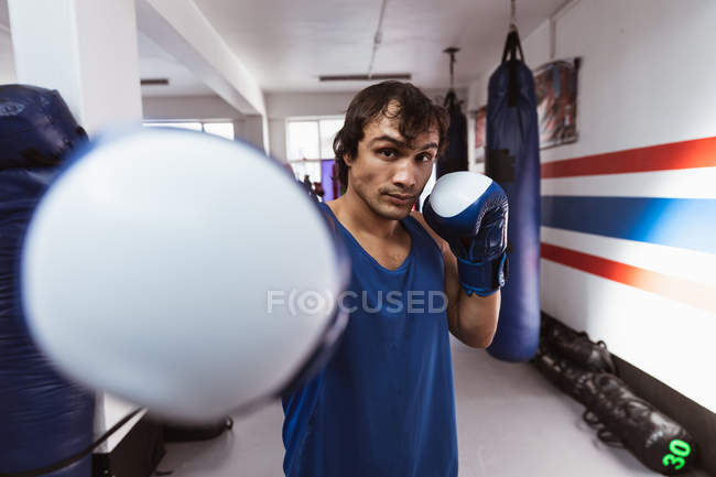 Vista frontal de cerca de un joven boxeador masculino de raza mixta en un gimnasio de boxeo golpeando a la cámara - foto de stock