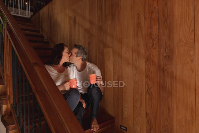 Vista frontal de um homem e mulher caucasiano de meia-idade sentado em uma escada em sua casa, segurando canecas de café e beijando — Fotografia de Stock