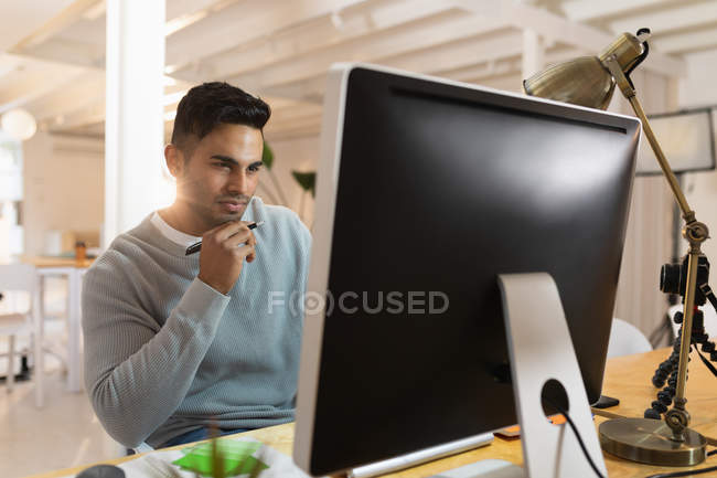 Вид спереду крупним планом молодий змішаний чоловік, що сидить за столом і дивиться на екран комп'ютера в творчому офісі — стокове фото