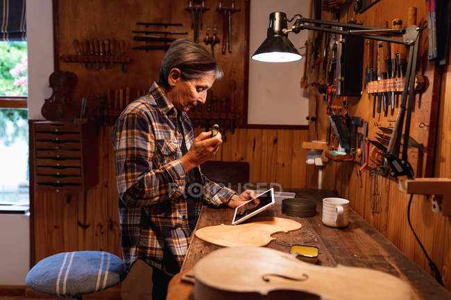 Vista lateral de un luthier femenino caucásico senior usando una tableta con herramientas colgando en la pared en el fondo - foto de stock