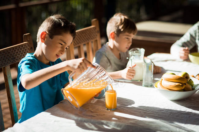 Vista lateral de cerca de dos niños caucásicos pre adolescentes sentados en una mesa disfrutando de un desayuno familiar en un jardín - foto de stock
