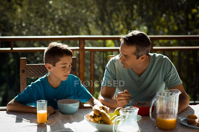 Vista frontal close-up de um homem caucasiano de meia-idade sentado em uma mesa com seu filho pré-adolescente, desfrutando de um café da manhã em família em um jardim — Fotografia de Stock