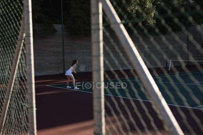 Vista lateral de uma jovem caucasiana jogando tênis, segurando uma raquete e esperando a bola vista através de uma cerca — Fotografia de Stock