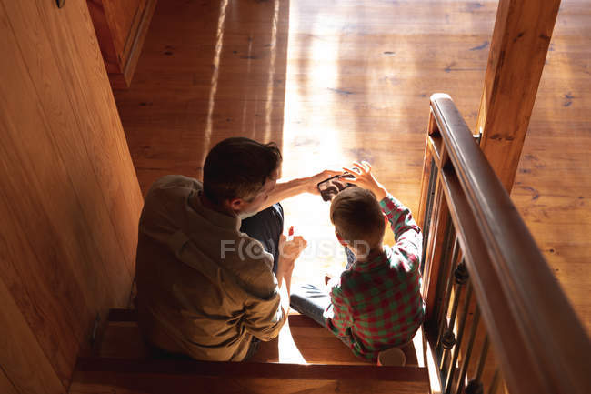 Visão traseira de um homem caucasiano de meia-idade e seu filho pré-adolescente sentado em uma escada em casa, usando um smartphone — Fotografia de Stock