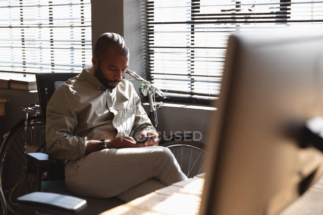 Vista frontal close-up de um jovem afro-americano sentado em uma mesa por uma janela usando um smartphone em um escritório criativo — Fotografia de Stock