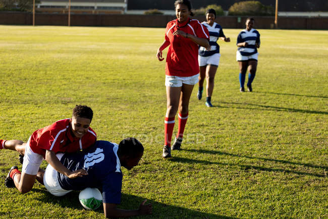 Вид спереду на групу молодих дорослих багатоетнічних гравців регбі під час матчу, три гравці бігають і один з м'ячем на землю — стокове фото