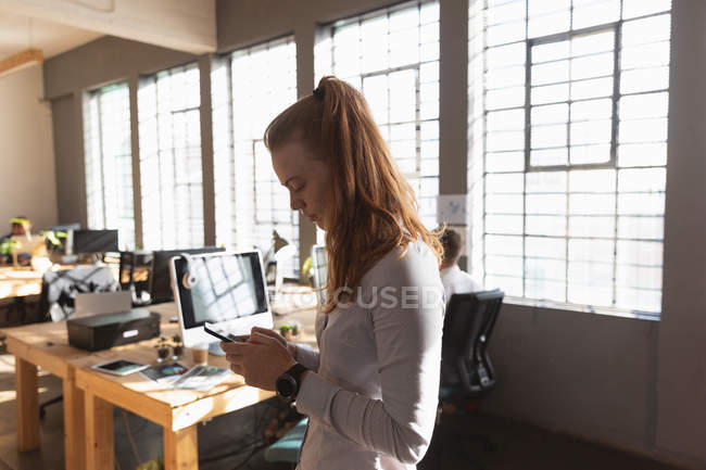 Vista lateral de perto de uma jovem caucasiana de pé usando um smartphone em um escritório criativo com mesas e computadores em segundo plano — Fotografia de Stock