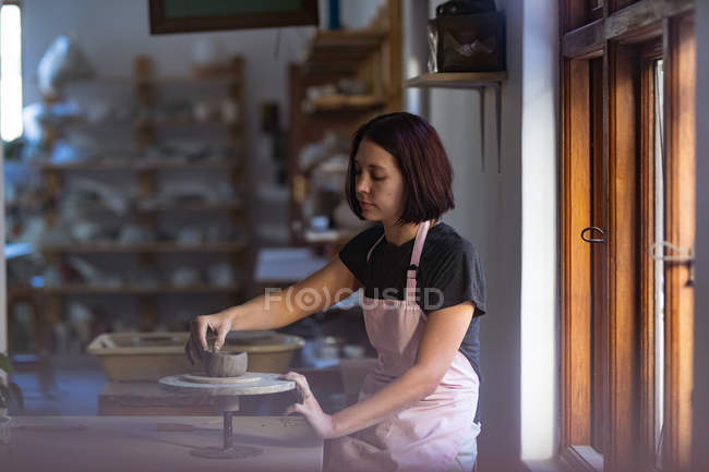 Vue latérale d'une jeune potière caucasienne assise à une table de travail devant une fenêtre, travaillant avec de l'argile sur une roue de baguage dans un atelier de poterie — Photo de stock