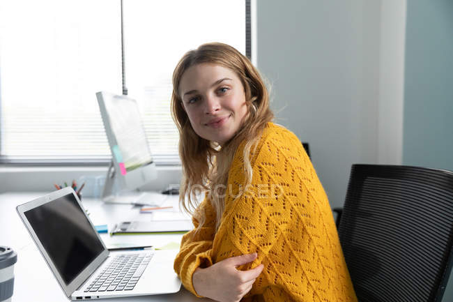 Портрет молодої кавказької жінки, яка користується ноутбуком і посміхається, сидячи за столом у сучасному офісі творчого бізнесу. — стокове фото