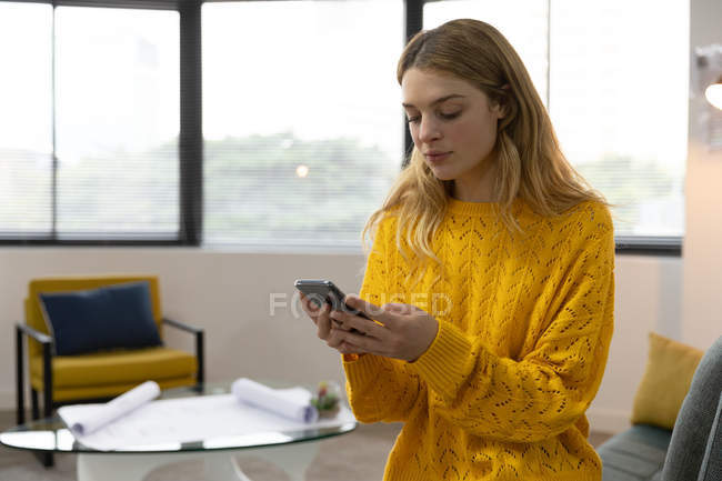 Vista frontal de cerca de una joven mujer caucásica usando un teléfono inteligente en la oficina moderna de un negocio creativo - foto de stock