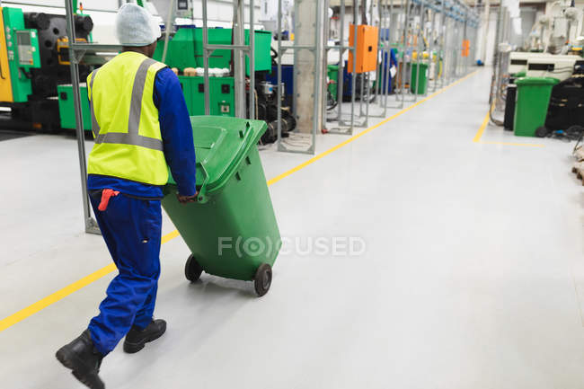 Задний план молодой афроамериканец, работающий на фабрике, везет зеленый мусорный бак через склад на перерабатывающем заводе — стоковое фото