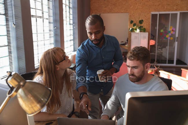 Vista frontal de cerca de un joven afroamericano de pie sosteniendo una taza de café hablando con una joven mujer caucásica y colegas masculinos sentados en un escritorio usando una computadora en una oficina creativa - foto de stock