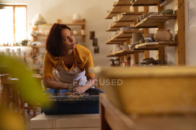 Вид на молодий Кавказький жіночий Поттер сидить і працює з глиною на гончарних колесо в гончарної студії, і дивлячись на бік посміхаючись, поки вона працює, з обладнанням на передньому плані — стокове фото