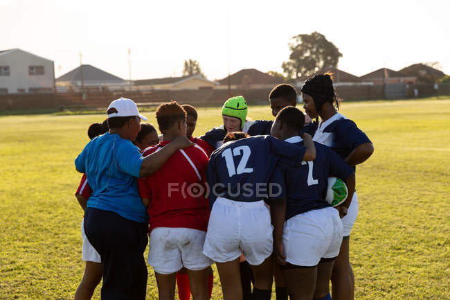 Visão traseira de jovens adultas multi-étnicas jogadoras de rugby do sexo feminino e sua raça mista de meia idade treinador em pé em um aconchego em um campo de rugby durante um jogo — Fotografia de Stock