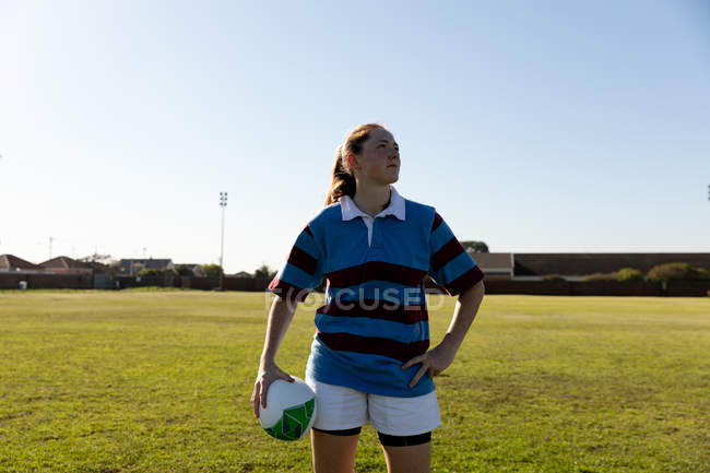 Вид спереди молодой белой женщины-регбистки, стоящей на регби, смотрящей в сторону с рукой на бедре и держащей регбийный мяч. — стоковое фото