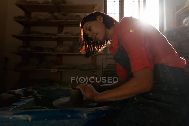 Vista laterale di una giovane ceramista caucasica sorridente seduta e che lavora con argilla su una ruota di vasai in uno studio di ceramica, retroilluminata dalla luce del sole — Foto stock