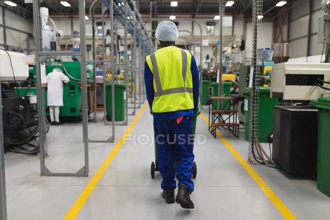Rear view close up of a young African American male factory worker wheeling a bin through a warehouse at a processing plant, the rear view of another factory worker operating a machine is visible in the background — Stock Photo