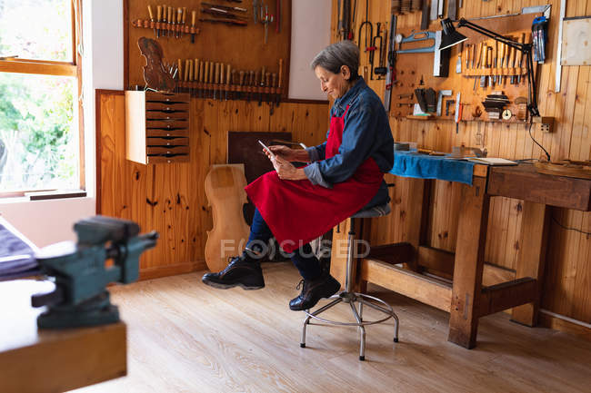 Вид збоку старших кавказьких жіночих luthier в майстерні за допомогою планшетного комп'ютера з інструментами висить на стіні у фоновому режимі — стокове фото