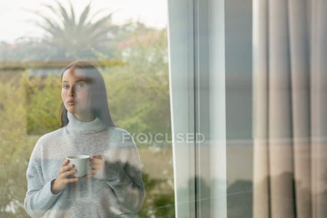 Front view of a young Caucasian brunette woman wearing a grey turtleneck sweater, standing on a balcony holding a cup of coffee, seen from inside, through window — Stock Photo
