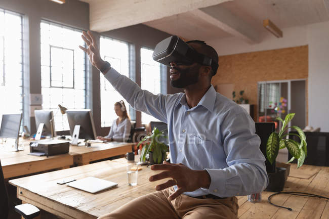 Side view close up of a young African American man sitting on a desk wearing a VR headset with his arms outstretched and hands out in a creative office, a female colleague working in the background — Stock Photo