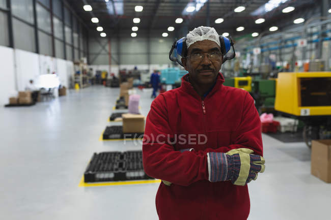 Porträt eines afrikanisch-amerikanischen Fabrikarbeiters mittleren Alters in Arbeitskleidung mit Ohrenschützern auf dem Kopf, der in einer Lagerhalle eines Verarbeitungsbetriebs steht und mit verschränkten Armen in die Kamera blickt — Stockfoto