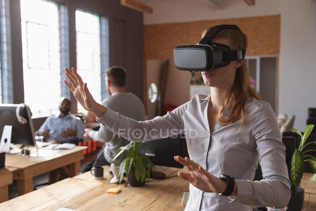 Vue de face gros plan d'une jeune femme caucasienne portant un casque VR debout avec les bras tendus et les mains dans un bureau créatif, avec des collègues travaillant en arrière-plan — Photo de stock