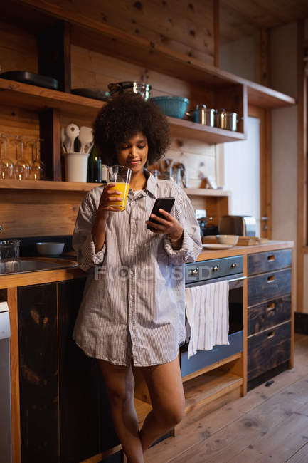 Nahaufnahme einer jungen Frau mit gemischter Rasse in einem Hemd, das mit einem Smartphone steht und zu Hause in ihrer Küche ein Glas Orangensaft hält — Stockfoto