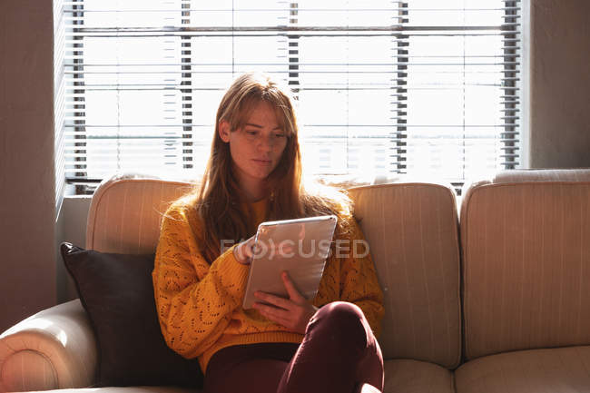 Vista frontal de perto de uma jovem caucasiana sentada em um sofá usando um tablet na área de estar de um escritório criativo, iluminada pela luz solar — Fotografia de Stock